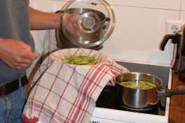 Edamame in a cooking pot on the stove. One person has poured a portion of edamame into a sieve covered with a kitchen cloth.